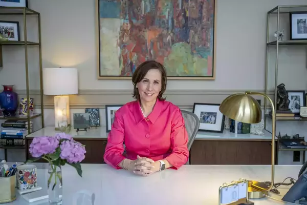 Ann Firth sits at her desk. There's a vase of blue hydrangeas on the desk, and a colorful abstract painting in the background.